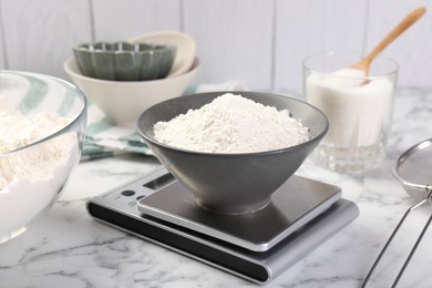 Photo of Digital kitchen scale with bowl of flour and other products on white marble table, closeup