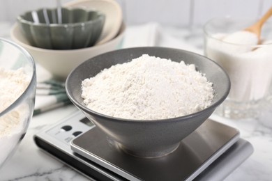 Photo of Digital kitchen scale with bowl of flour and other products on white marble table, closeup