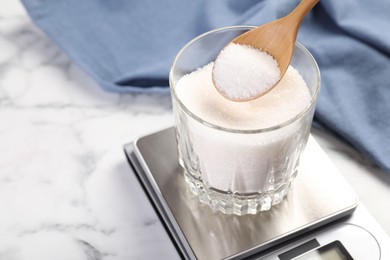 Photo of Putting sugar into glass on digital kitchen scale at white marble table, closeup. Space for text