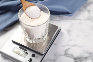Photo of Putting sugar into glass on digital kitchen scale at white marble table, closeup. Space for text