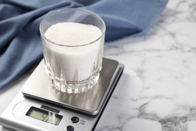 Photo of Digital kitchen scale with glass of sugar on white marble table, closeup