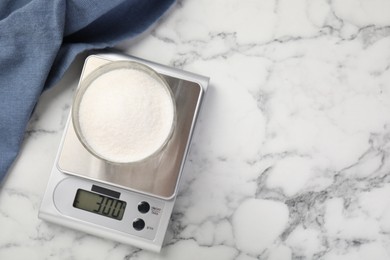 Photo of Digital kitchen scale with glass of sugar on white marble table, top view. Space for text