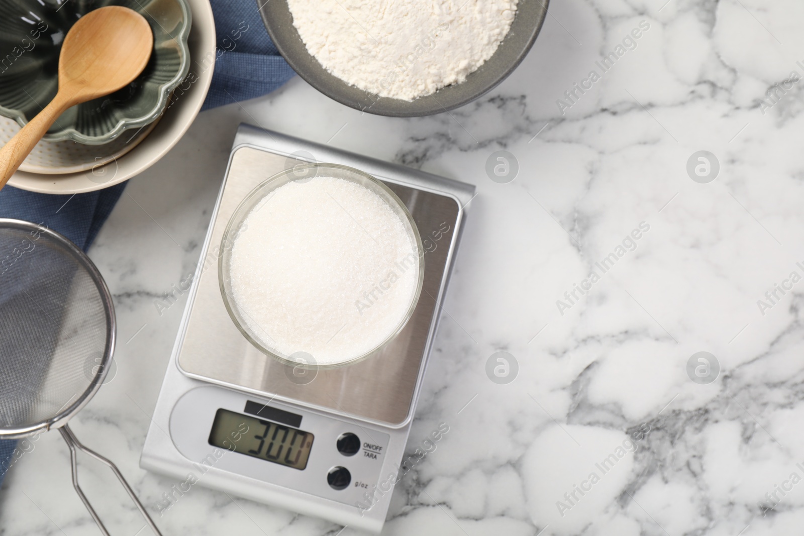 Photo of Digital kitchen scale with glass of sugar and other products on white marble table, flat lay. Space for text