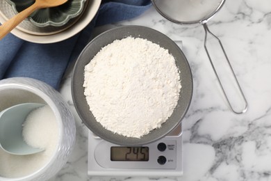 Photo of Digital kitchen scale with bowl of flour and other products on white marble table, flat lay