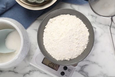 Photo of Digital kitchen scale with bowl of flour and other products on white marble table, flat lay