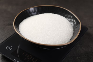 Photo of Digital kitchen scale with bowl of sugar on grey table, closeup