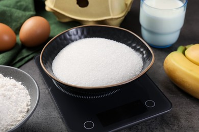 Photo of Digital kitchen scale with bowl of sugar and other products on grey table, closeup