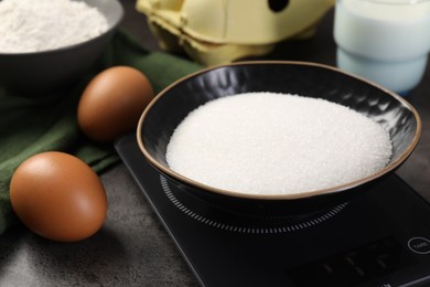 Photo of Digital kitchen scale with bowl of sugar and other products on grey table, closeup