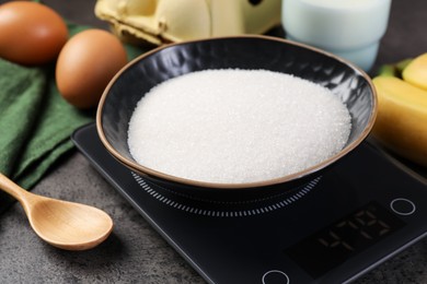Photo of Digital kitchen scale with bowl of sugar and other products on grey table, closeup