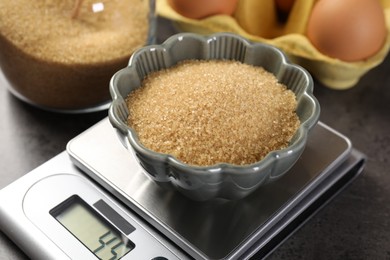 Photo of Digital kitchen scale with bowl of brown sugar and other products on grey table, closeup