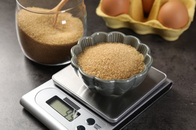 Photo of Digital kitchen scale with bowl of brown sugar and other products on grey table, closeup