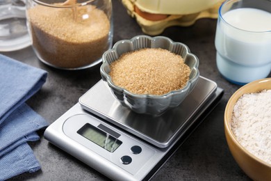 Photo of Digital kitchen scale with bowl of brown sugar and other products on grey table, closeup