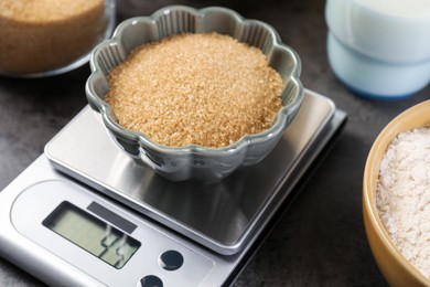 Photo of Digital kitchen scale with bowl of brown sugar and other products on grey table, closeup