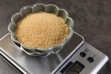 Photo of Digital kitchen scale with bowl of brown sugar on grey table, closeup