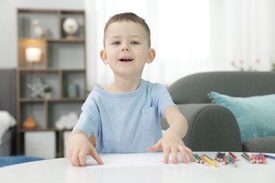 Photo of Cute little boy with sheet of paper and colorful pencils at table indoors