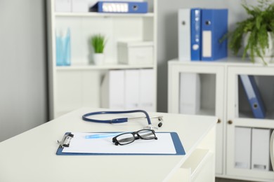 Photo of Doctor's workplace. Stethoscope, clipboard, pen and glasses on white table in medical office