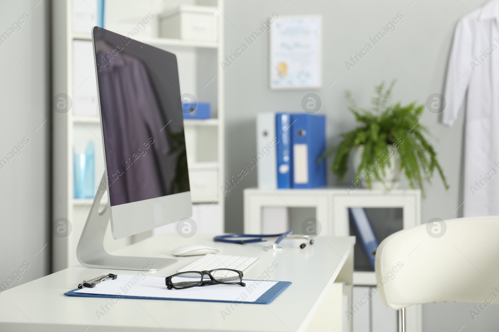 Doctor's workplace. Computer monitor, clipboard, stethoscope and glasses on white table in medical office Photo of Doctor's workplace. Computer monitor, clipboard, stethoscope and glasses on white table in medical office