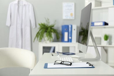 Photo of Doctor's workplace. Computer monitor, clipboard, stethoscope and glasses on white table in medical office