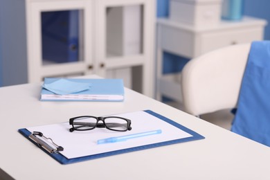 Photo of Doctor's workplace. Clipboard, other stationery and glasses on white table in medical office