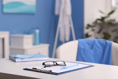 Photo of Doctor's workplace. Clipboard, other stationery and glasses on white table in medical office