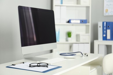 Photo of Doctor's workplace. Computer monitor, clipboard, stethoscope and glasses on white table in medical office