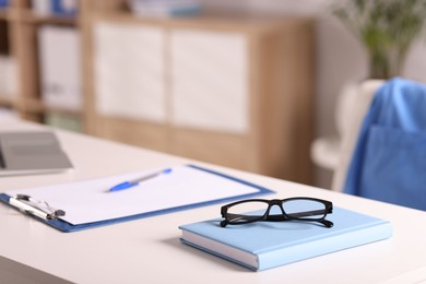 Photo of Doctor's workplace. Notebook, glasses, clipboard and pen on white table in medical office, closeup. Space for text