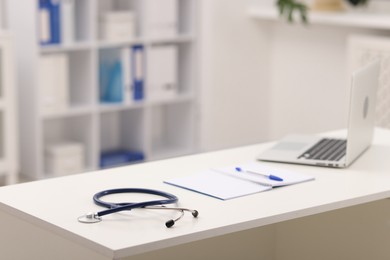 Photo of Doctor's workplace. Stethoscope, laptop, open notebook and pen on white table in medical office, space for text