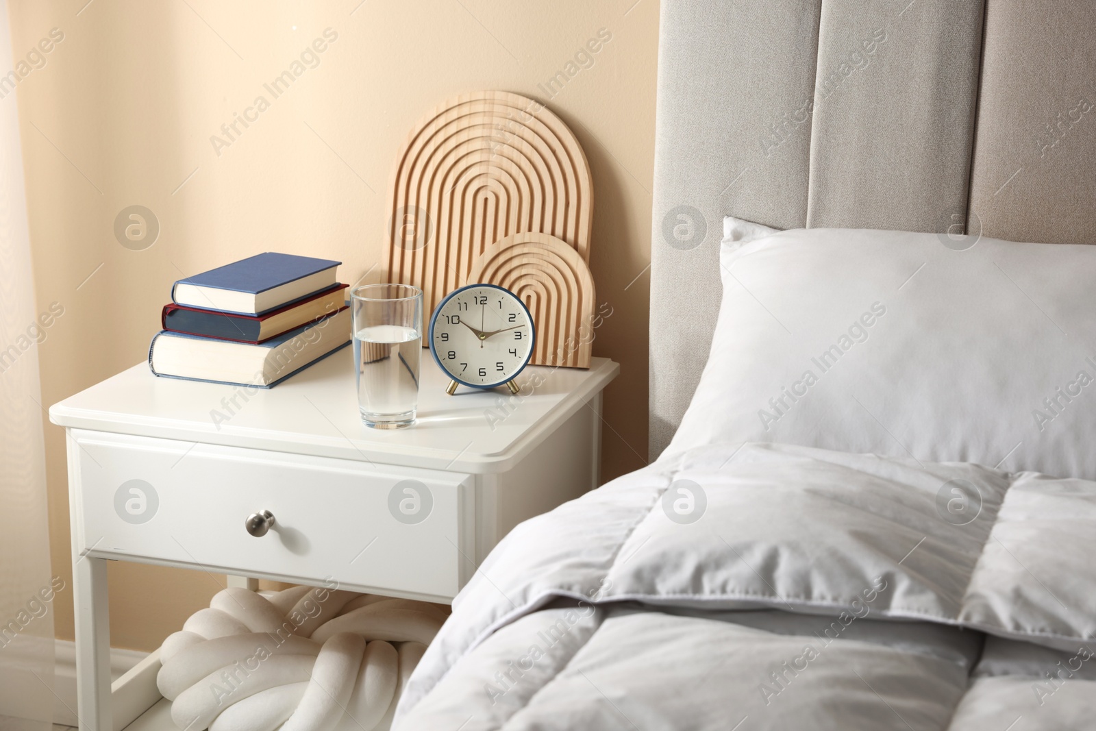 Bedside table with books, decor, glass of water and alarm clock near bed in room Photo of Bedside table with books, decor, glass of water and alarm clock near bed in room