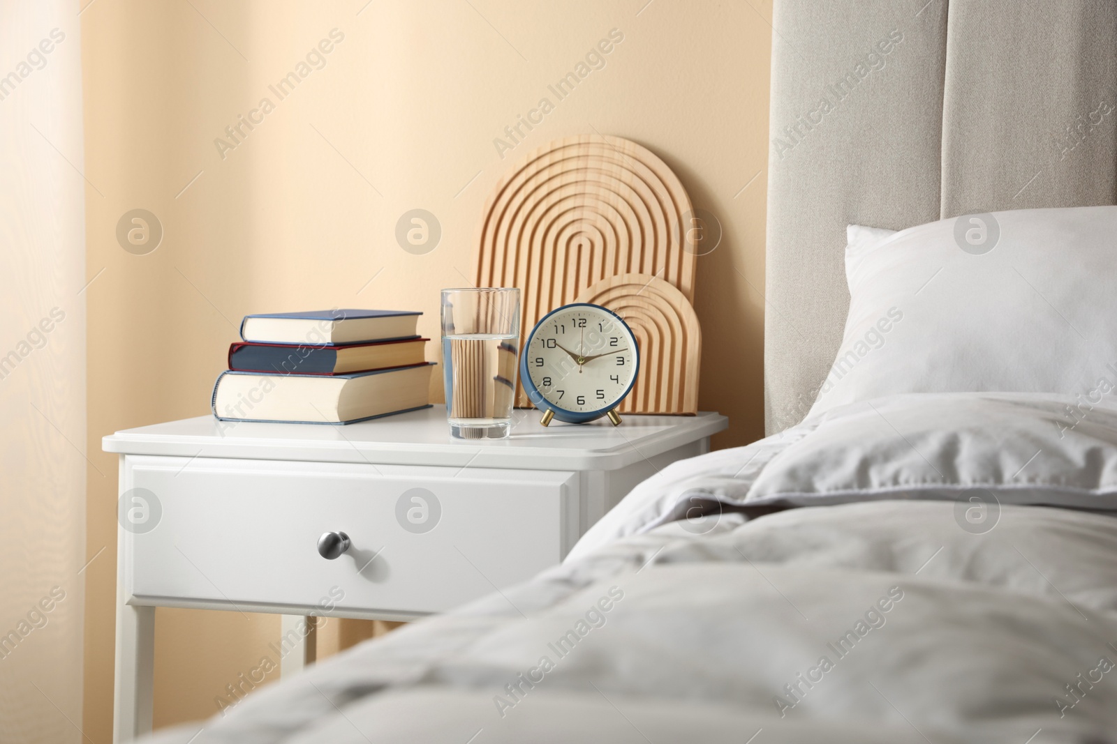 Bedside table with books, decor, glass of water and alarm clock near bed in room Photo of Bedside table with books, decor, glass of water and alarm clock near bed in room