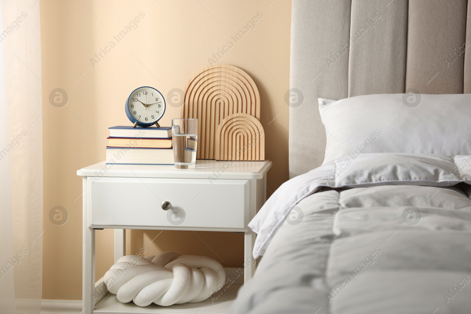 Bedside table with books, decor, glass of water and alarm clock near bed in room Photo of Bedside table with books, decor, glass of water and alarm clock near bed in room