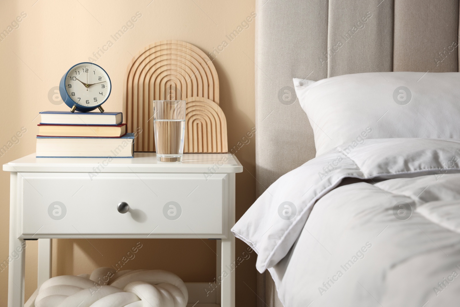 Bedside table with books, decor, glass of water and alarm clock near bed in room Photo of Bedside table with books, decor, glass of water and alarm clock near bed in room