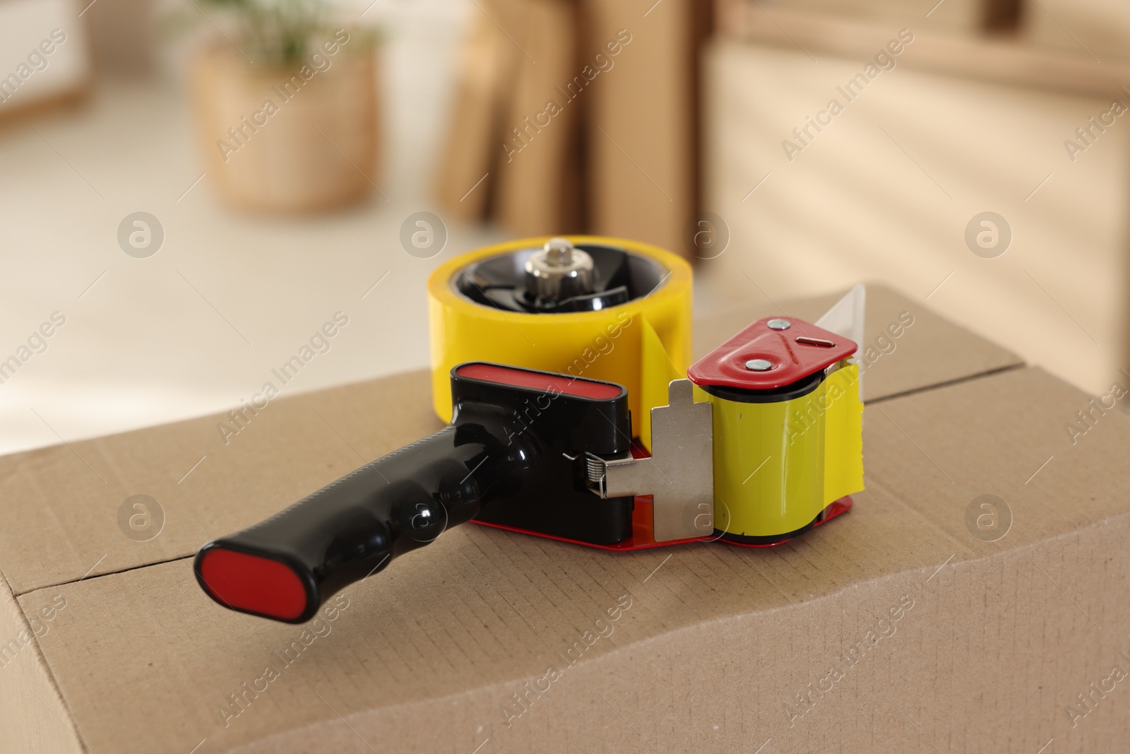 Photo of Dispenser with adhesive tape on cardboard box indoors, closeup