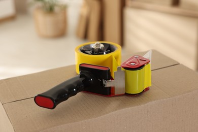 Photo of Dispenser with adhesive tape on cardboard box indoors, closeup
