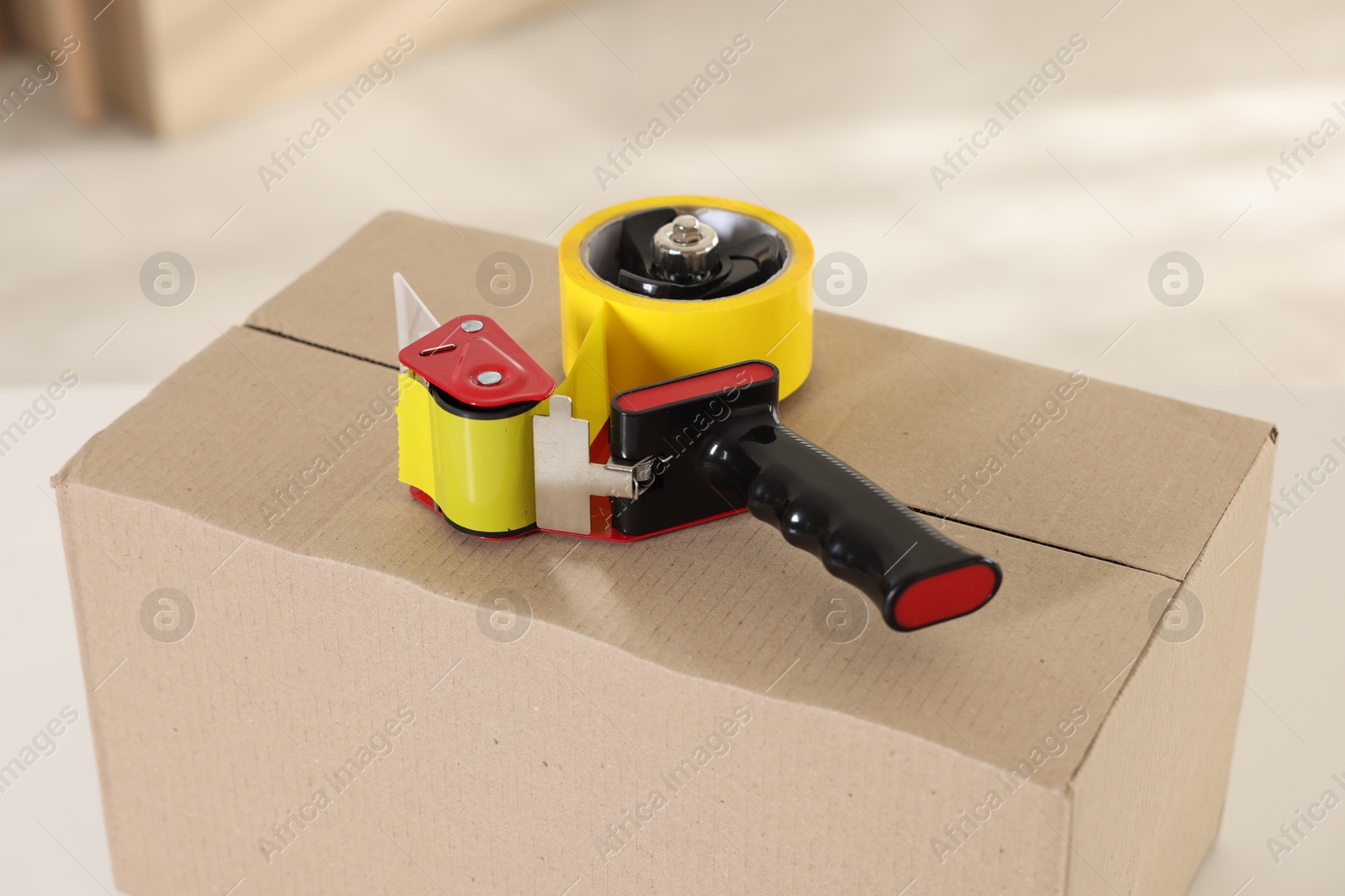 Photo of Dispenser with adhesive tape on cardboard box indoors, closeup