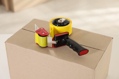 Photo of Dispenser with adhesive tape on cardboard box indoors, closeup