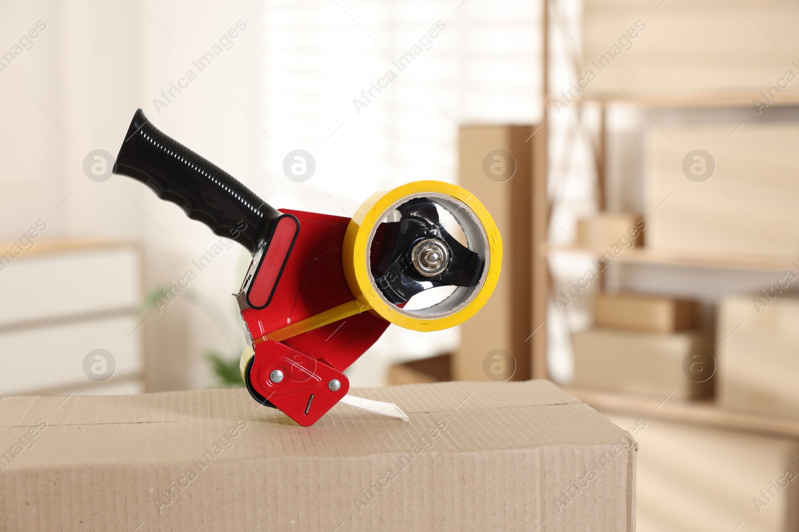 Photo of Dispenser with adhesive tape on cardboard box indoors, closeup