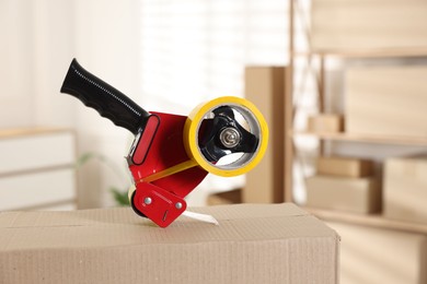 Photo of Dispenser with adhesive tape on cardboard box indoors, closeup
