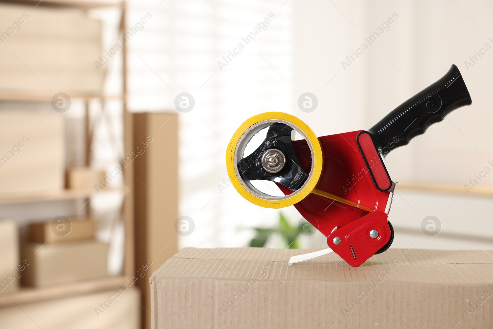 Photo of Dispenser with adhesive tape on cardboard box indoors, closeup