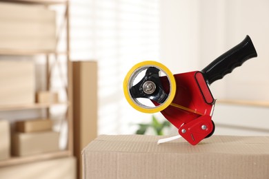 Photo of Dispenser with adhesive tape on cardboard box indoors, closeup