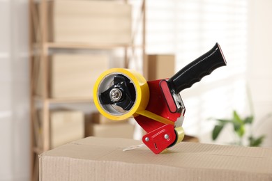 Photo of Dispenser with adhesive tape on cardboard box indoors, closeup