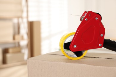 Photo of Dispenser with adhesive tape on cardboard box indoors, closeup