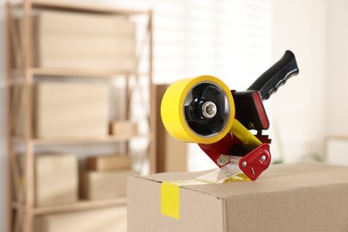 Photo of Dispenser with adhesive tape on cardboard box indoors, closeup