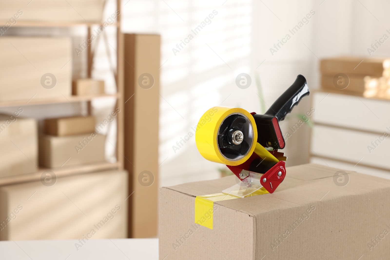 Photo of Dispenser with adhesive tape on cardboard box indoors, closeup