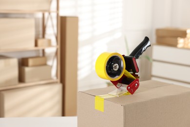 Photo of Dispenser with adhesive tape on cardboard box indoors, closeup