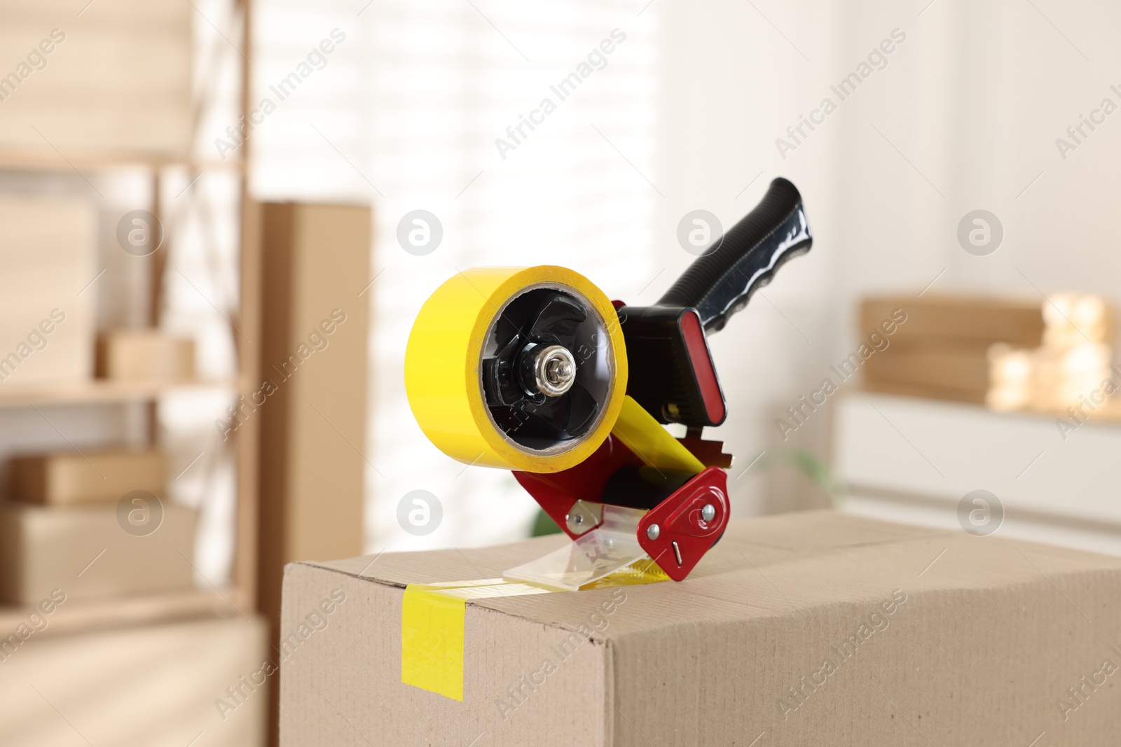 Photo of Dispenser with adhesive tape on cardboard box indoors, closeup
