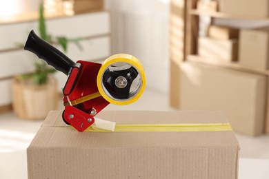 Photo of Dispenser with adhesive tape on cardboard box indoors, closeup