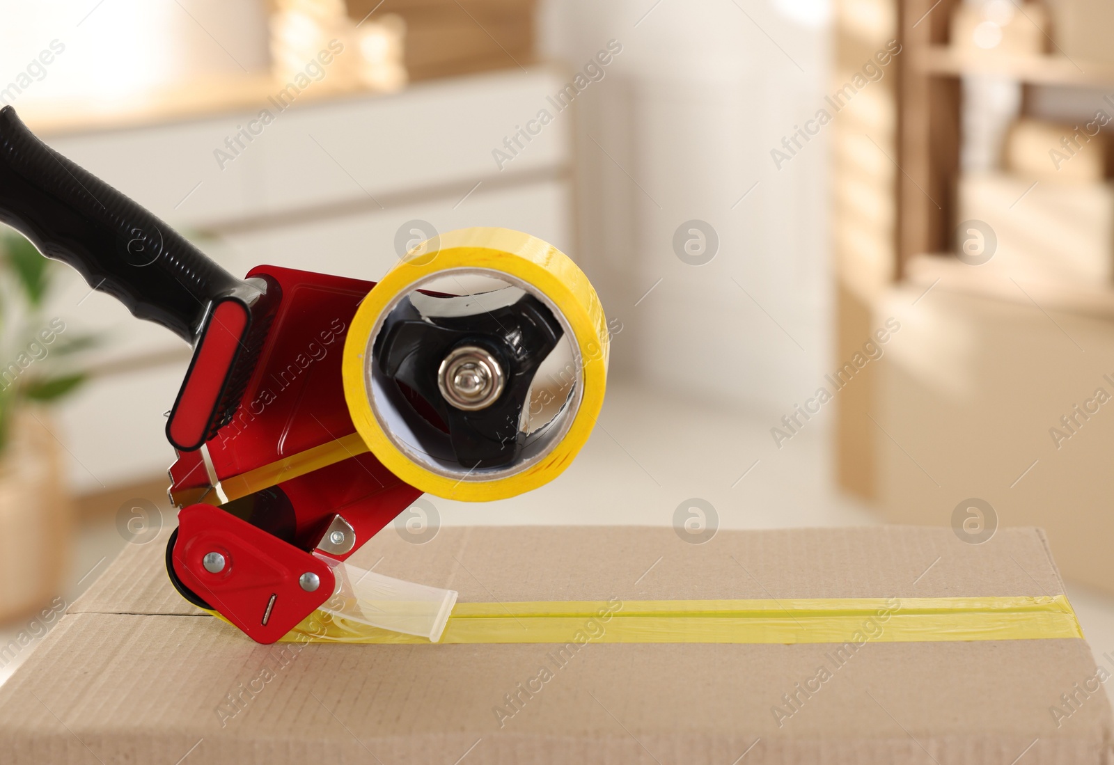Photo of Dispenser with adhesive tape on cardboard box indoors, closeup