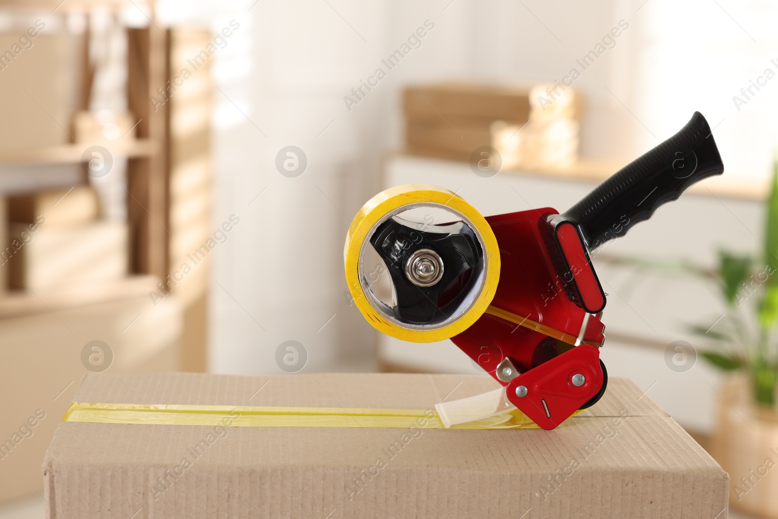 Photo of Dispenser with adhesive tape on cardboard box indoors, closeup