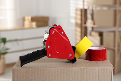 Photo of Dispenser with adhesive tape rolls on cardboard box indoors, closeup