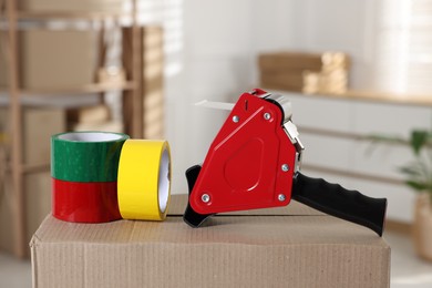 Photo of Dispenser with adhesive tape rolls on cardboard box indoors, closeup